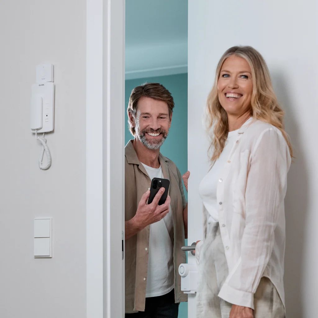 Couple at the door using the Nuki Smart Lock, with a smartphone in hand and an intercom visible on the wall.